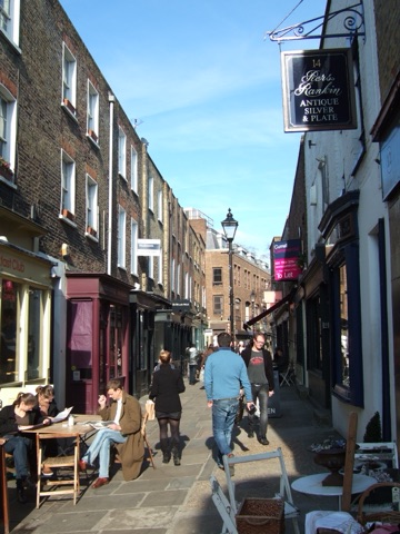 Camden Passage in low winter sun: a narrow flagged lane of Georgian brick, an antique dealer's hanging sign over the shopfronts, people walking down the middle and two women reading at a café table