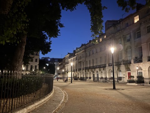 Fitzroy Square at dusk, the lamps lit along the paved centre and the Georgian terrace glowing on the right