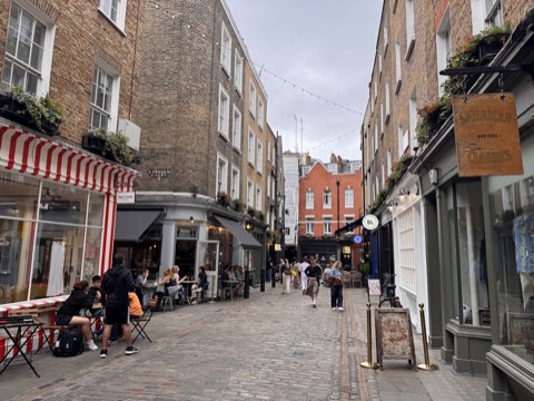 Newburgh Street's cobbles with a striped awning on the left, small shopfronts on both sides and people walking in the middle