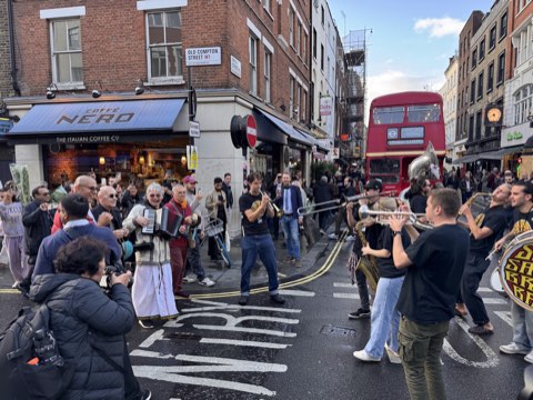 A brass band playing in the road at the Wardour Street corner of Old Compton Street, a red bus held up behind them