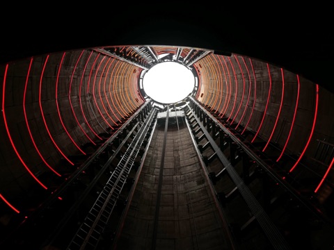 Looking straight up the inside of the chimney from the lift: ribbed concrete walls lit by red strips running the full height, the lift rails and maintenance ladders converging towards the top, and a circle of white sky at the end of it