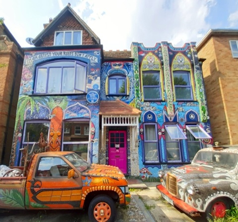 The front of a terraced house covered entirely in mosaic: I'M AN ARTIST YOUR RULES DON'T APPLY spelled in tiles across the top, sugar skulls and patterned arches worked around the windows, a bright pink front door under a small tiled porch, and two mosaic-covered vehicles at the kerb - an orange pick-up truck and a silver saloon.