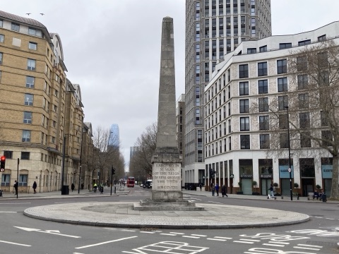 A stone obelisk on a paved traffic island at the centre of St George's Circus, roads and modern blocks running away on either side, with the dedication to Brass Crosby carved on its plinth.