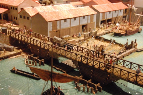 A museum model of the Roman bridge: a timber bridge on piles crossing the river, with a covered cart, soldiers and traders on it, red-roofed warehouses along the quay behind and ships moored at the wharf
