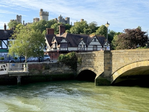 The River Arun flowing under a stone bridge, a timber-framed building on the far bank and the towers of Arundel Castle above the trees behind.