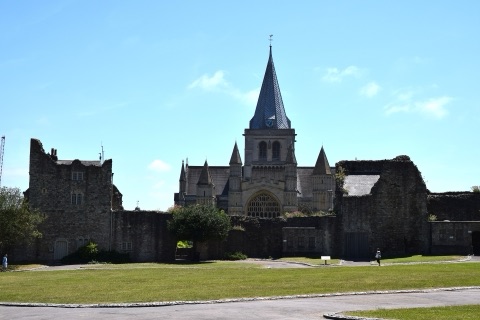 Rochester Cathedral's Norman west front and its slender green-blue spire seen over the ruined walls of the castle bailey, across a lawn on a bright day.