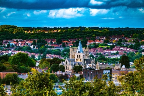 Rochester's cathedral spire and the square keep of the castle among the red roofs of the town, under a dark bank of cloud, with wooded hills behind.