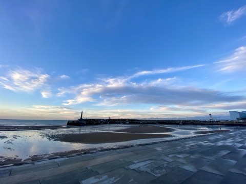 Margate's wide sands at low tide with the stone harbour arm and its small lighthouse across the bay, under a big blue sky with streaks of cloud.
