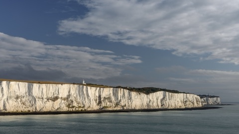 A long wall of white chalk cliffs above a calm sea under a sky of grey and white cloud, with a small white lighthouse on the green clifftop.