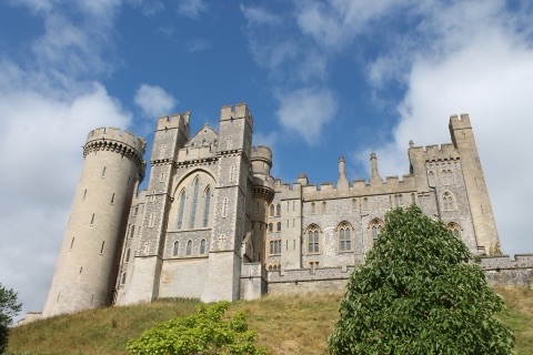 Arundel Castle's round keep, gothic towers and battlemented ranges of grey stone rising above a grass bank against a blue sky with white clouds.