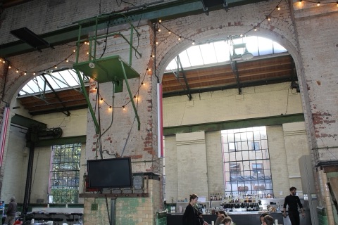 The turbine hall as a restaurant: tall brick arches with strings of bulbs, a green iron gantry and old machinery overhead, a bar along the far wall and diners at tables below.