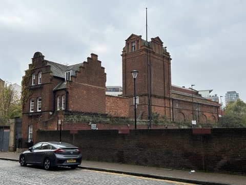 The power station from Wapping Wall: a red-brick engine house with a Dutch gable and a square tower with a pedimented top, behind a long brick wall on a cobbled street, under a grey sky.