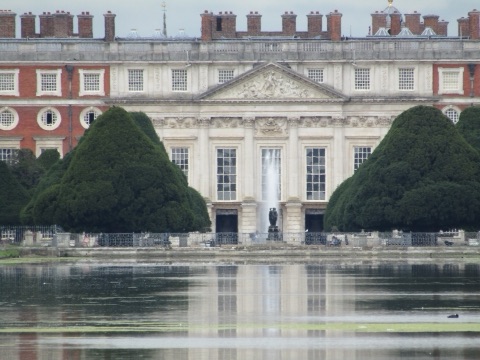The east front of Hampton Court Palace across the Long Water: a pedimented centre of pale stone between red-brick wings, a fountain playing in front, and two huge clipped yews either side of it.