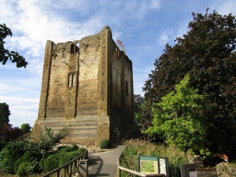 The square stone keep of Guildford Castle on its mound, with a flag on top, a path leading up past trees and a notice board on a summer day.