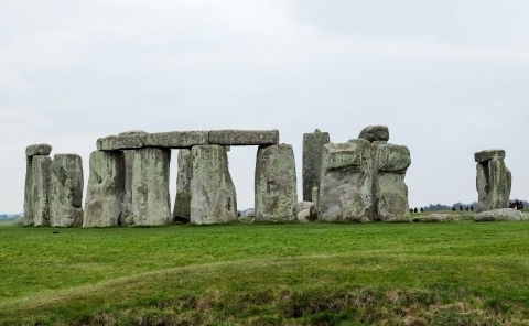 The standing stones of Stonehenge under a grey sky: the tall trilithons with their lintels, fallen stones around them, on a green plain with a few visitors at the far side.