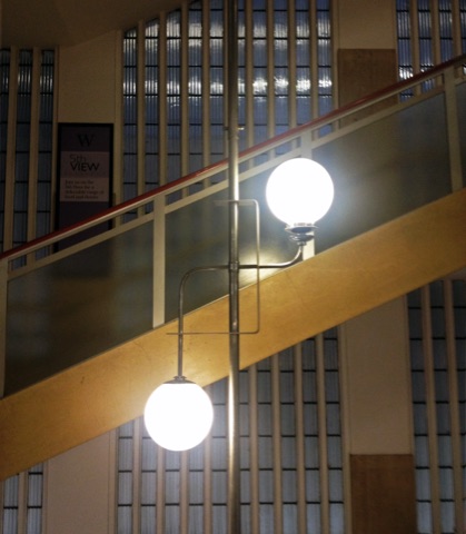 A chrome stair lamp with two white glass globes on a slender pole, against a sweeping staircase with a glass balustrade and a wall of fluted glass panels.