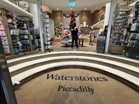 The shop's entrance from the pavement: curved travertine steps with Waterstones Piccadilly set into the floor in dark letters, glass doors either side, and tables of books, a Christmas tree and a spiral staircase inside.