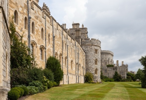 A long stone range of the castle with rows of gothic windows running away to a round tower, a strip of lawn and shrubs along its foot.