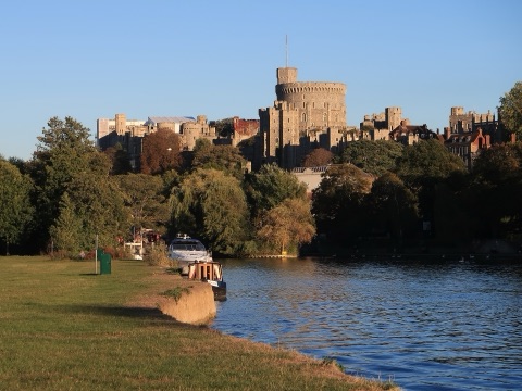Windsor Castle's grey Round Tower and battlemented walls on a hill above the Thames, seen across the river from a grass bank in evening light, with two boats moored below the trees.