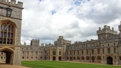 A wide lawn enclosed by long stone ranges with rows of tall windows and battlemented towers, under a cloudy sky.